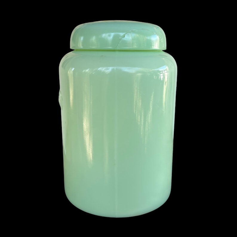 May include: A pale green jar with a lid, cylindrical in shape. The jar has a smooth, shiny surface and a rounded bottom. The lid fits securely, creating a complete seal. The jar is probably made of glass or ceramic.