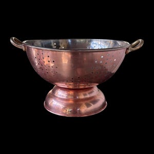 Puede incluir: Un colador de cobre pulido que presenta un cuenco ancho y redondeado con numerosos orificios de drenaje, una base de pedestal resistente y dos asas laterales curvas. La superficie interior es de color plata metálica, contrastando con el acabado.