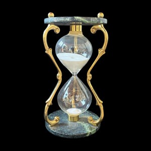 May include: An hourglass featuring a clear glass bulb filled with white sand, held by a polished gold-tone metal frame with ornate curved supports. The top and bottom bases are crafted from dark green marble with visible veining patterns.