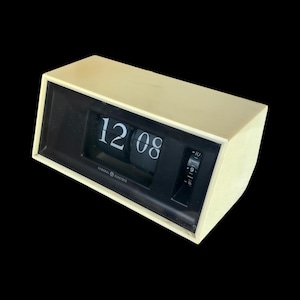 Puede incluir: Un reloj de solapa General Electric vintage en una caja color crema con una cara negra. El reloj muestra la hora 12:08 en números blancos. El reloj tiene un diseño retro y un pequeño dial de ajuste en el lateral.