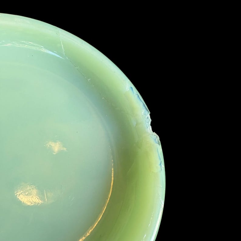 May include: Close-up of a pale green glass bowl with a cracked rim. The bowl has a smooth, glossy surface and a slight opalescent quality. The background is solid black.