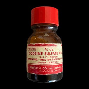 Könnte beinhalten: Eine kleine bernsteinfarbene Glasflasche mit einem leuchtend roten Schraubverschluss. Das weiße Etikett trägt den roten Text POISON, 1/3 oz., CODEINE SULFATE MERCK, U.S.P.-POWDER, WARNING-May be habit forming, OPIUM DERIVATIVE und MERCK & CO. Inc., Rahway, N.J.