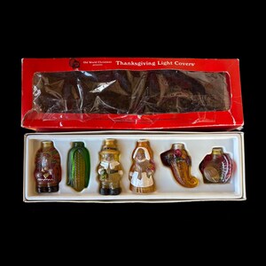 May include: A set of Thanksgiving light covers in a red box. The set includes six glass light covers shaped like a Native American, corn, a pilgrim, a woman, a cornucopia, and a pumpkin. The box reads "Thanksgiving Light Covers."