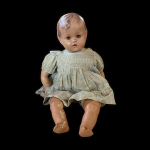 Könnte beinhalten: Eine antike Babypuppe mit bemaltem Gesicht, braunen Augen und hellbraunem Haar. Die Puppe trägt ein hellblaues Kleid mit Blumenmuster. Die Beine der Puppe weisen Gebrauchsspuren auf, wobei etwas Farbe abgeblättert ist.