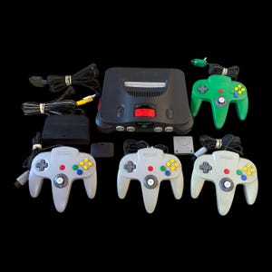 Op de afbeelding: Een antracietgrijze Nintendo 64-spelcomputer met een rode uitbreidingsklep, één groene controller, drie lichtgrijze controllers, een zwarte voeding, een grijze geheugenkaart en diverse zwarte stroom- en AV-kabels met witte kabelbinders.