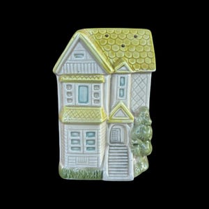 May include: A ceramic house-shaped salt or pepper shaker. The house is white with a yellow-green roof and trim. It has multiple windows with blue accents, a front door, and a small tree on the side. The shaker has two holes on the roof.