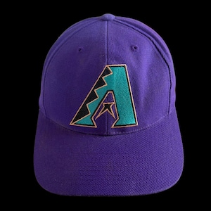 Puede incluir: Una gorra de béisbol morada con una letra A bordada en la parte frontal. La letra es de color verde azulado con un contorno negro y un borde cobrizo. La gorra tiene una corona estructurada y una visera curva, hecha de tela morada.