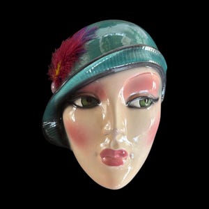 May include: A ceramic face sculpture with a teal cloche hat adorned with a red and purple feather. The face has green eyes, pink blush, and glossy pink lips. The background is black.