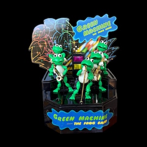 May include: A Green Machine The Frog Band musical display featuring four green frog figures on a black stage. The frogs are playing instruments including a saxophone and guitar. The display includes blue signage with the text Green Machine The Frog Band.