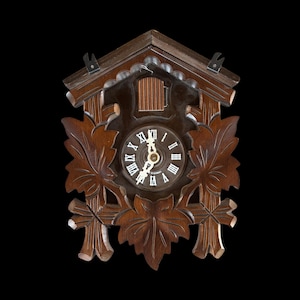 Puede incluir: Un reloj de cuco de madera marrón oscuro con detalles de hojas talladas. La esfera del reloj tiene números romanos y agujas doradas. El reloj está sobre un fondo negro.