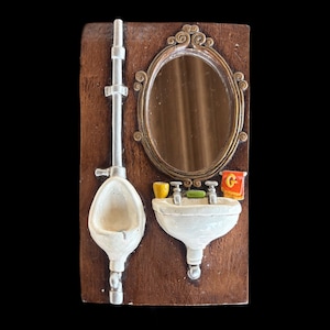 May include: A miniature bathroom scene with a white urinal, sink, and mirror. The mirror has an ornate gold frame. A small yellow cup and a red towel with a gold "G" are on the sink. The background is a dark brown.