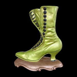 May include: A lime green decorative boot with black buttons and laces, set on a brown, ornate base. The vintage-style boot features a pointed toe and a small heel, with a glossy finish. The boot is a decorative item.