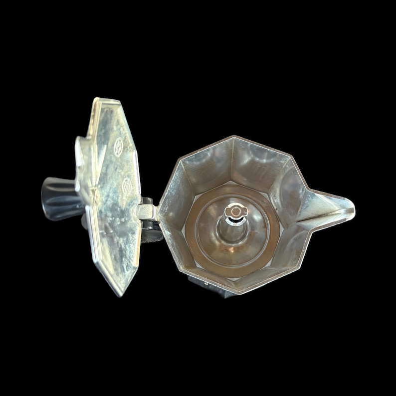 May include: A silver-colored stovetop espresso maker, open to reveal the interior components. The lid is open, showing the filter basket and the spout. The handle is black.