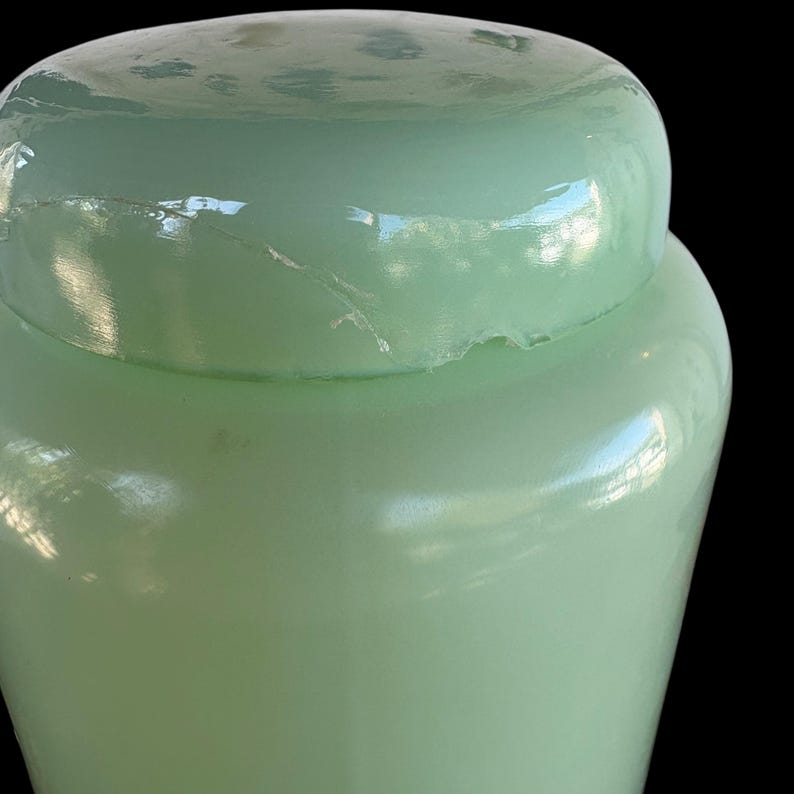 May include: A pale green glass jar with a rounded lid. The jar has a smooth, glossy surface and a small crack near the lid. The jar is set against a black background.