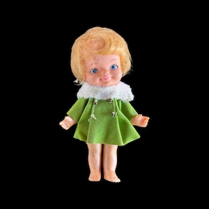 Puede incluir: Muñeca vintage con cabello rubio y ojos azules, vestida con un abrigo verde con cuello de piel blanco. La muñeca tiene una expresión dulce y está sobre un fondo negro.