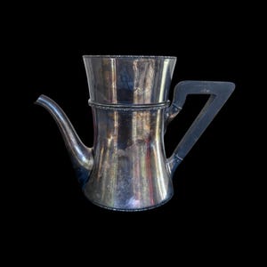 Pode incluir: Uma cafeteira prateada com bico, alça e uma seção superior separada. A cafeteira tem um design clássico com um bico curvo e uma alça escura. O fundo é preto.