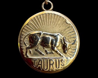 May include: A round gold-toned pendant featuring a raised relief of a bull in the center with a sunburst pattern in the background. The word TAURUS is embossed in capital letters at the bottom. The pendant hangs from a gold-toned chain.