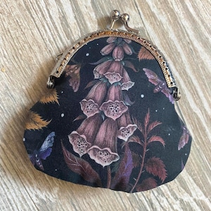 Könnte beinhalten: Eine kleine, dunkel gefärbte Geldbörse mit Metallverschluss. Die Geldbörse hat ein Blumendesign mit lila und rosa Blumen, Blättern und Schmetterlingen. Der Metallverschluss ist silberfarben.