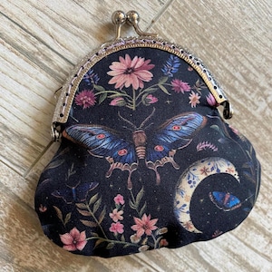 Pode incluir: Uma bolsa de moedas preta com fecho prateado, com uma borboleta de asas azuis e detalhes florais. O design inclui flores rosa e roxas, uma lua crescente e outros elementos botânicos.