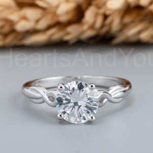 May include: A silver-toned ring featuring a single round, clear, faceted stone held by four prongs. The band has a twisted, leaf-like design on both sides of the center stone. The text HeartsAndYou is visible in the background behind the ring.