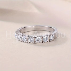 May include: A silver band ring featuring a row of seven round, clear gemstones. The ring is set on a light-colored surface, showcasing the sparkle of the stones. The ring is a piece of jewelry.