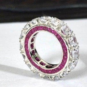 May include: A silver eternity ring featuring a central band of square-cut vibrant pink rubies flanked by two outer rows of round-cut clear white stones. The band has a metallic finish and a circular design with stones set around the entire circumference.