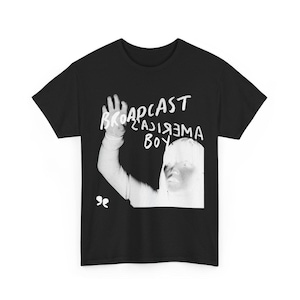 Peut inclure: T-shirt noir avec un graphisme blanc. Le graphisme présente le texte "BROADCAST AMERICA BOY" et une image stylisée d'une personne avec un bras levé. Le t-shirt a un col rond et des manches courtes.