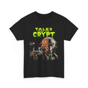 May include: A black short-sleeved t-shirt featuring a graphic print of the Crypt Keeper from Tales from the Crypt. The text TALES FROM THE CRYPT is printed in a bright green, dripping font above the character, who is shown with wispy hair.
