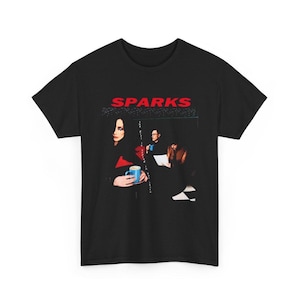 Op de afbeelding: Zwart T-shirt met de naam van de band "SPARKS" in rode letters. Daaronder een afbeelding van drie mensen, waarvan één een blauwe mok vasthoudt. Het T-shirt heeft een ronde hals.