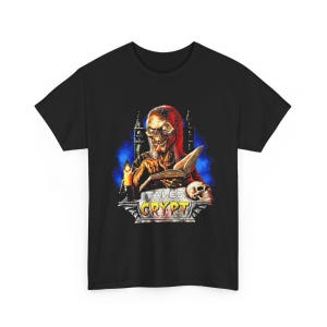 Peut inclure: Un t-shirt noir avec une impression graphique du Crypt Keeper de Tales from the Crypt. Le personnage est représenté en train de lire un livre avec un crâne et une bougie à proximité, sur un fond bleu avec le texte Tales from the Crypt en dessous.