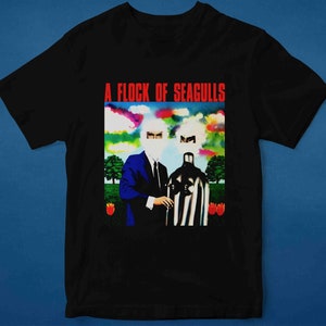 Op de afbeelding: Zwart T-shirt met een kleurrijke afbeelding van twee figuren met bedekte gezichten. De afbeelding heeft de titel "A Flock of Seagulls".