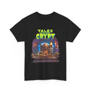 May include: A black short-sleeved t-shirt featuring a graphic print of the Crypt Keeper from Tales from the Crypt. The design shows the character holding a candle over an open book, with the text TALES FROM THE CRYPT in bright green letters.