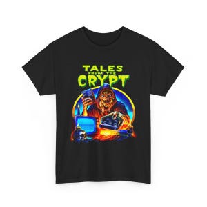 Puede incluir: Una camiseta negra de manga corta con un estampado gráfico de una figura esquelética que sostiene un mando a distancia y un libro. El texto "TALES FROM THE CRYPT" aparece en verde sobre la figura, con un televisor, velas y una calavera.