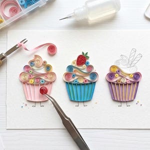 May include: A white paper sheet featuring three paper quilling cupcakes in pink, blue, and purple. The cupcakes are decorated with colorful coiled paper swirls and a red strawberry. Crafting tools include a slotted tool, tweezers, and glue.