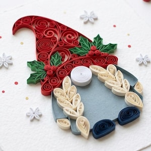 May include: A paper quilling art piece featuring a gnome with a red coiled hat, green holly leaves, and red berries. The gnome has a white coiled nose, cream-colored leaf-shaped beard accents, and dark blue feet, surrounded by small white paper snowflakes.