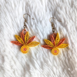 May include: A pair of handmade earrings featuring a floral design. The earrings are crafted from rolled paper in shades of yellow, orange, and peach. They have gold-colored ear wires and a circular base, set against a white, fluffy background.