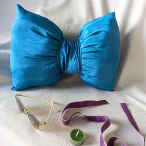 Peut inclure: Un grand coussin turquoise en forme de nœud papillon, fait d'un tissu brillant. Le coussin est au centre, avec des lunettes de soleil à monture dorée, un ruban violet et une petite bougie verte à proximité. Les objets sont disposés sur une surface blanche.