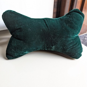 May include: A dark green, bow-shaped pillow made of a soft, velvety material. The pillow is designed with rounded edges and a slight curve in the center. It is placed on a white surface.