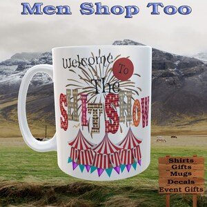 May include: White ceramic mug with the text "Welcome To The SHITSHOW" in a circus-themed design. The mug features a handle and a colorful illustration of circus tents. The background shows a mountain range and a grassy field.