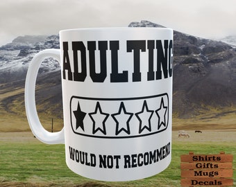 May include: White ceramic mug with the words "ADULTING WOULD NOT RECOMMEND" in black, along with a star rating graphic. The mug has a curved handle and is set against a scenic background. The text "Men Shop Too" is at the top.