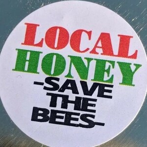 Peut inclure: Un autocollant rond blanc avec le texte "LOCAL HONEY - SAVE THE BEES" en lettres rouges, vertes et noires.