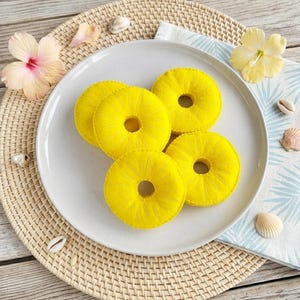 Peut inclure: Cinq tranches d'ananas jaunes en feutre, percées d'un trou central, sont disposées sur une assiette blanche. L'assiette repose sur un dessous de plat tressé, avec une serviette à motifs bleus et blancs, des coquillages et des fleurs d'hibiscus.