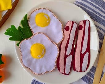May include: Two felt fried eggs with bright yellow yolks and white edges, two strips of felt bacon in dark red and light pink, and a green felt lettuce leaf arranged on a white plate. The food items are crafted from soft fabric materials.