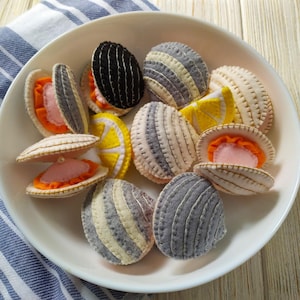 May include: A collection of handcrafted felt clams and lemon slices arranged in a white bowl. The clams feature stitched details in shades of grey, black, and cream, with some open to reveal orange and pink interiors, one containing a small white pearl.