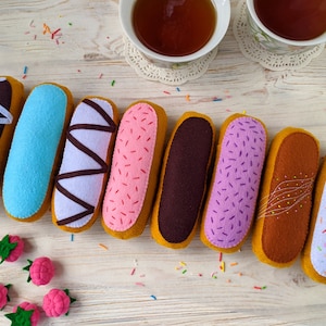 May include: Eight felt eclair toys in various colors including brown, blue, lavender, pink, and white, decorated with felt icing, zig-zag patterns, and sprinkles. Four small pink felt raspberries with green leaves are placed near the eclairs.