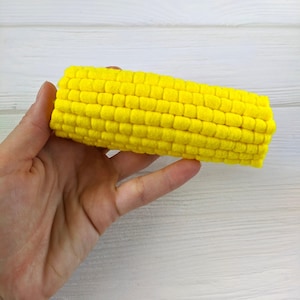 May include: A bright yellow felt corn cob toy featuring a textured surface with individual rounded kernels arranged in a grid pattern. The object is cylindrical in shape and crafted from soft fabric material, mimicking the appearance of fresh corn.