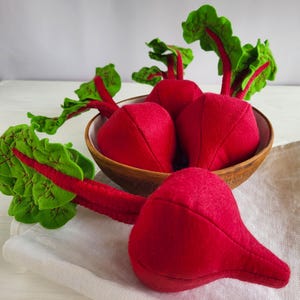 May include: A wooden bowl filled with plush, red beets with green and red leaves. One beet rests outside the bowl on a white cloth. The beets have a soft, felt-like texture, and the leaves have detailed stitching.