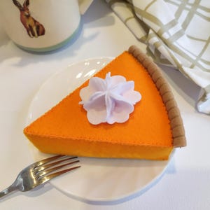 May include: A felt slice of orange pumpkin pie on a white plate with a silver fork. The pie features a brown crust edge and a white dollop of whipped cream on top. The stitching is visible along the edges of the orange felt fabric.