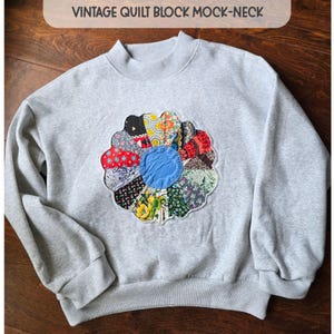 May include: A light gray mock-neck sweatshirt featuring a large, circular, multi-colored floral quilt block appliqué on the chest. The appliqué consists of various patterned fabric segments surrounding a solid blue center. Text reads VINTAGE QUILT BLOCK MOCK-NECK.