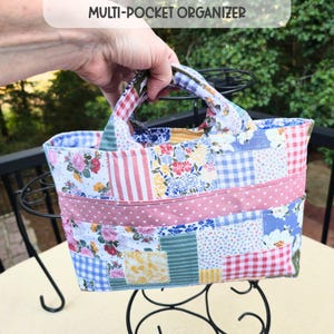 May include: A multi-pocket organizer bag featuring a patchwork design with various floral, gingham, and striped patterns in shades of pink, blue, yellow, and green. The fabric bag includes a horizontal pink band with white hearts and two top handles.